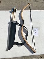 Top view of the wooden recurve bow, black quiver with carrying strap, and six wooden arrows with colored fletching on a white table.