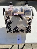 Full view of the bath gift kit placed outdoors on a white surface, showing the fabric basket with black leaf designs holding a transparent container with various bath items inside.