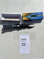 Photo of folding knife unfolded showing two blades, black metal handles with cut-out patterns, original blue branded box, and black sheath.