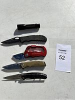 Top view of four folded knives, red utility cutter, and small black tool on white surface.