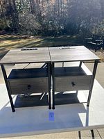 Pair of matching end tables shown side by side outdoors on a white table. Both have a dark wood finish top with black metal legs and a power outlet panel on top.