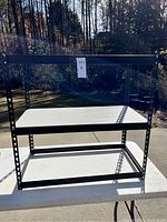 Front view of black metal shelving unit with white metal shelves outdoors on a white table in natural light.