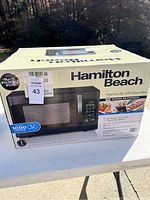 Boxed Hamilton Beach microwave oven in black stainless steel with product image and features on the box.