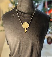 Necklace with circular patterned disk pendant and wing drop hanging on black fabric torso display.