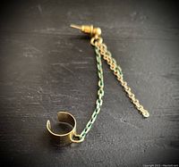 Close-up photo of the gold plated earring/cuff showing the brass stud and two chains, one gold plated and one green faceted, against a dark background.
