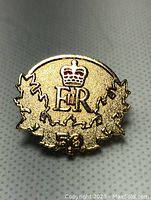 Close-up photo of a gold-tone lapel pin with Queen Elizabeth II's crowned insignia (E II R) surrounded by maple leaves and the number '50' for the Golden Jubilee 2002.