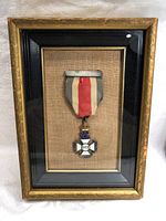 Full view of framed World War II era King George VI cross medal enclosed in a gold frame with black inner borders. Medal hangs from a red, white, and blue ribbon on a burlap-textured background.