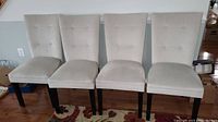 Front view of 4 matching beige cushioned dining chairs with button tufting and dark wooden legs arranged side by side.