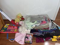 Children's clothing laid out on floor showing dresses, shirts, pants, plush animals, toy hockey sticks, and a colorful box of shapes and colors.