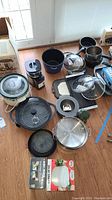 Photo showing assortment of cookware and kitchen appliances including pots, pans, chopper, salad spinner, and slow cooker components on wooden floor.
