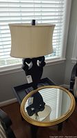 Full view of black sculptural table lamp with beige shade next to round mirror with gold frame on table