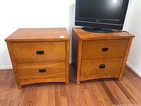 Two matching wooden nightstands placed side-by-side, both with two drawers and black metal handles. One nightstand has a TV on top for scale (not included).