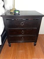 Front view of one black wooden nightstand showing three drawers with metal ring pull handles, placed on a wooden floor near a bed.