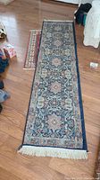 Photo of long narrow runner rug with intricate floral patterns in blue, cream, muted red, and beige colors, placed on wooden floor.