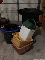 Front view of all items grouped including large trash can, blue wash basin with liner, watering can, white and yellow storage bins, and peach hamper basket.