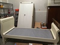 Full view of assembled trundle bed frame with mattress removed, cream color, stored indoor, no drawers.