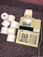 Beige Sharp adding machine beside multiple paper rolls indicating it includes several printer paper rolls.