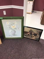 Two framed art prints leaning against a wall: the larger floral print with white lilies in a green frame, and a smaller signed rustic building scene print in a black frame.