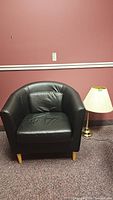 Black leather tub chair with light wood legs and matching pillow against pink wall and carpet. Partially visible brass table lamp with cream shade beside chair.