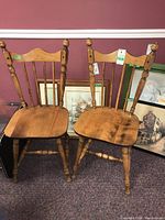 Two wooden dining chairs with spindled backrests, turned legs, and a natural wood finish. Chairs show signs of light wear and usage.