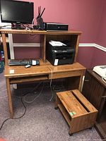 Composite wooden desk with upper shelving attached, showing a computer monitor, desktop PC, keyboard, printer, and wired computer mouse on and around the desk. A wheeled wooden printer stand is positioned in front of the desk.