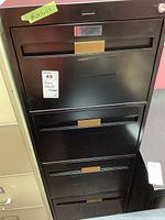 Front view of the black four drawer filing cabinet showing the lock and handles, with a yellow paper label indicating dimensions on top left.