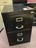 Front view of black metal two drawer filing cabinet showing handles, label holders, and visible wear