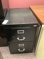Photo of a black metal two drawer filing cabinet with visible wear on top surface and metal handles on drawers.