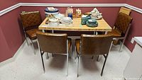 Full view of the rectangular kitchen table with an extendable leaf, topped with various kitchenware items, surrounded by four brown vinyl padded chairs with metal legs.