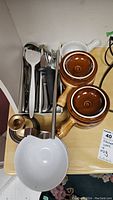 Photo of kitchenware set including white ladle, spatula, stainless silverware, brown ovenproof serving plates, and copper bowl.