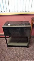 Front view of black metal file cabinet showing wear and scratches on top surface, lid closed, standing on carpet.
