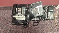 Five Norstar office phones grouped together on carpet floor, showing multiple line keys, coiled cords, and numeric keypads.