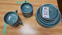Six turquoise-green cups stacked and arranged next to a stack of saucers and plates, showing overall condition and design.