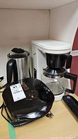 Photo showing the George Foreman electric grill, Hamilton Beach electric kettle, and Proctor Silex coffee maker on a shelf.