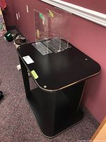 Front angled view of a black composite table with rounded corners and three clear acrylic holders on top, showing surface and shelving.