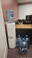 Full view of white water cooler with large water bottle on top, four additional empty water bottles with blue caps on floor beside it.