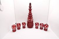 Full lot showing the red glass decanter with stopper centered among six small matching cordial glasses, all decorated with Christmas etchings.