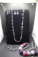 Photo of a black display board showing a long beaded necklace with small beads in pink, purple, white, and some silver tones; matching earrings; a chunky beaded necklace with large round beads in pink, purple, and gray; and a circular gray beaded bracelet.