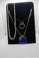 Three necklaces displayed on black velvet: hollow silver-coloured beads necklace, silver chain with double ring pendant, and silver chain with blue pendant.
