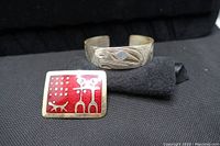 Photo of red enamel brooch with native figure and animal design beside a silver bracelet with native style engraving and blue stone.
