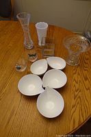 Photo showing mixed lot of glass vases, white glass vase, clear pressed glass, paintings on glasses, and white shaped bowls on wood table.