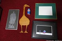 Top-down photo showing two framed art prints, a wooden goose decor, and an empty green and gold frame with a soda can for size reference on a red carpet