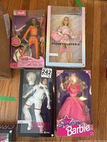 Four Barbie dolls in original packaging on floor: Samantha Cristoforetti astronaut Barbie, Superstar Barbie in pink gown, Cool Lookz Barbie in orange outfit, and Stoney Clover Barbie in pink pajama set.