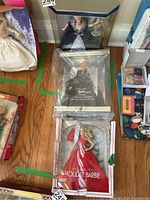 Three Barbie collector dolls displayed on a wooden floor in original packaging: one dressed in red and white, one in black and silver, and another in off-white.