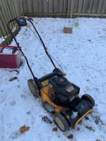 Side angle view of the yellow and black Cub Cadet SC 700h gas lawn mower in snow, showing handle, wheels, and engine area