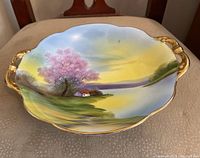 Top view of the larger porcelain serving tray showing hand-painted landscape scene with pink tree, lake, house, yellow sky, and gold handles and trim.