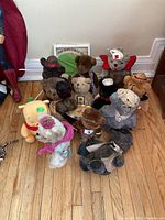 Front view of multiple assorted plush bears grouped together on wooden floor, including Winnie the Pooh and more bears wearing hats and scarves.