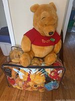 Large Winnie The Pooh plush bear sitting on top of a clear bag containing multiple smaller plush toys of Pooh characters.