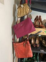 Front view of the three purses hanging on shelf; shows pink Guess purse, orange-brown Fossil purse, and gold mesh vintage purse