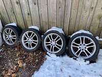 Four tire and rim assemblies leaning against a wooden fence with snow and leaves on the ground